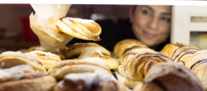 Delicious array of freshly baked pastries and cakes on display with a smiling baker in the background at Mad Hatter Cafe & Bakeshop in Durham