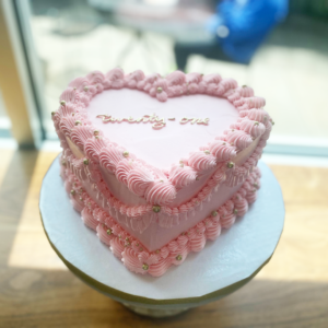 A heart-shaped cake with pink frosting, adorned with elegant piped decorations and the word "twenty-one" written on top, displayed on a white cake stand.