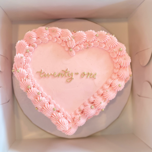 Heart-shaped cake with pink frosting and "twenty-one" written on top, inside a white cake box